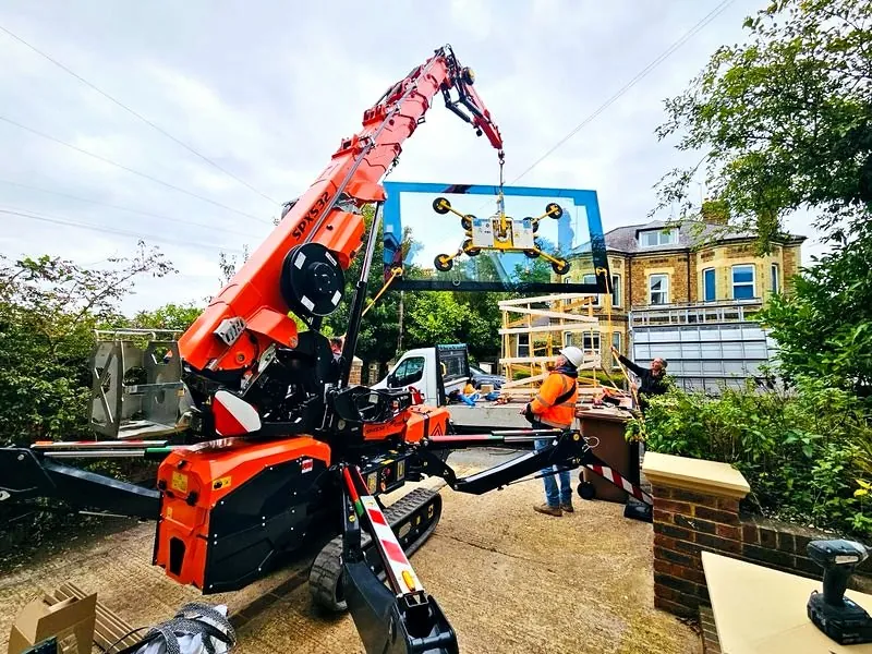 Orange Jekko SPX532 spider crane lifting a large glass pane with a vacuum lifter on a residential driveway
